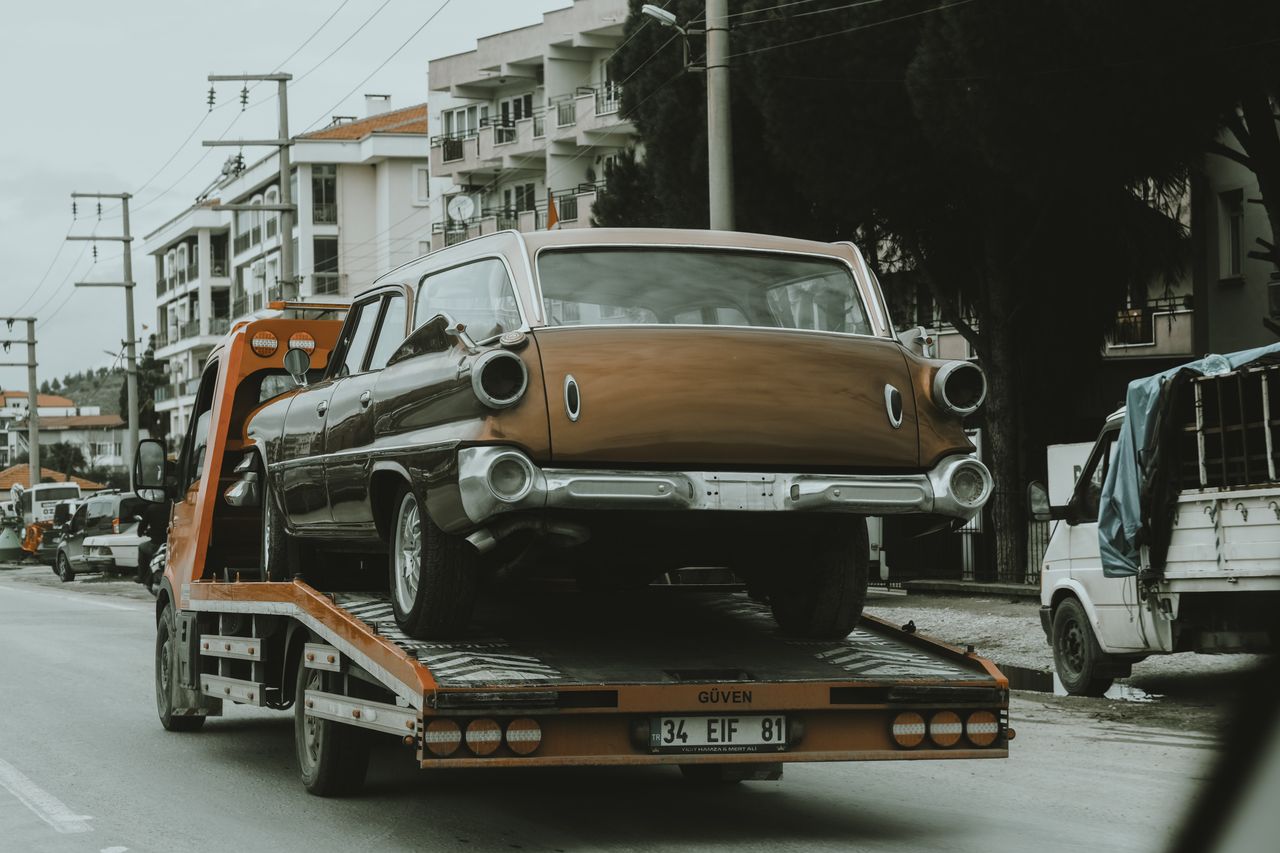 Home A vintage car loaded on a tow truck in a city street, surrounded by modern buildings.