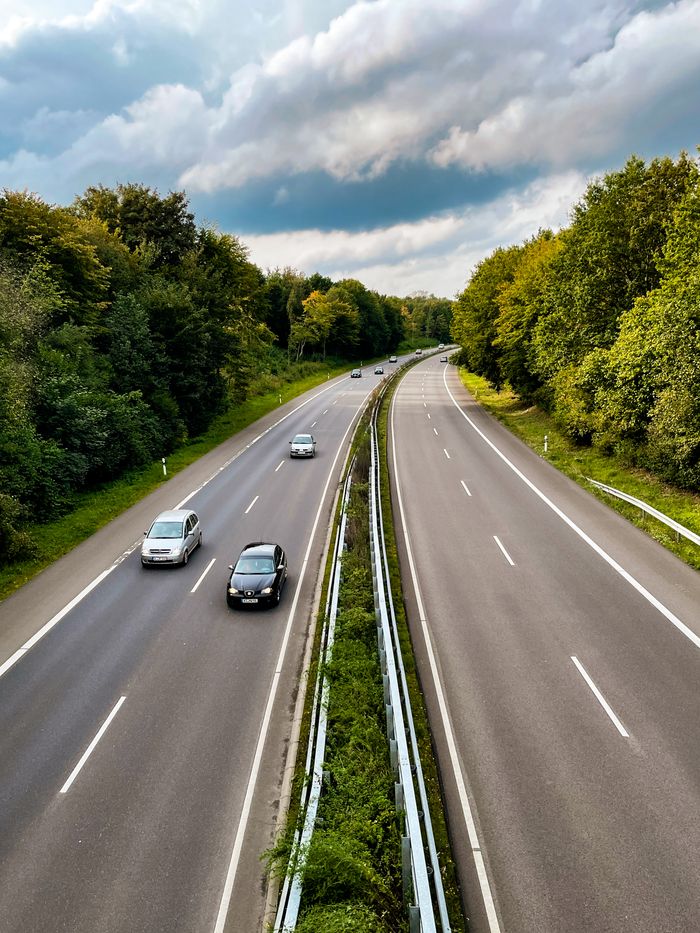 Home A peaceful highway scene with cars on an open road, flanked by lush green trees, under a cloudy sky.